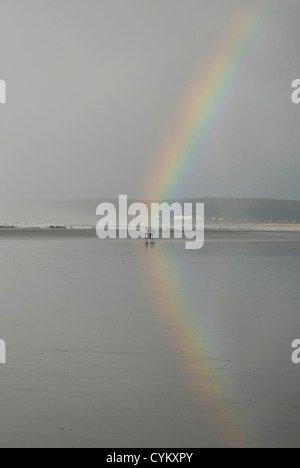 Un arc-en-ciel reflétées dans le sable humide sur la plage à Westward Ho ! North Devon après de fortes pluies. Les surfeurs se diriger vers la mer. Banque D'Images