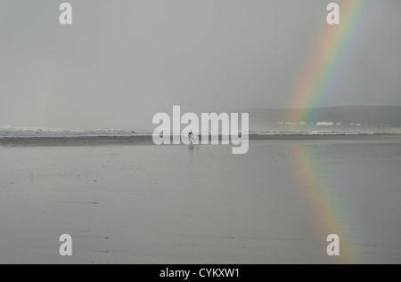 Un arc-en-ciel reflétées dans le sable humide sur la plage à Westward Ho ! North Devon après de fortes pluies. Les surfeurs se diriger vers la mer. Banque D'Images