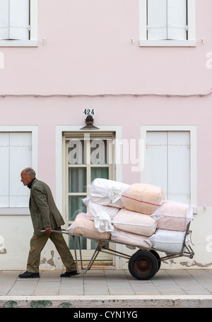 Un vieil homme tirant une charrette chargée de sacs blancs et roses marchant devant une maison rose Banque D'Images