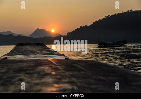 Bateaux en bois sur le fleuve Mékong au coucher du soleil Luang Prabang Laos // LUANG PRABANG, Laos — le fleuve Mékong coule devant cette ville classée au patrimoine mondial de l'UNESCO tandis que des bateaux traditionnels en bois traversent la voie navigable au loin. La lumière dorée filtre à travers la brume atmosphérique au coucher du soleil, créant des silhouettes des montagnes environnantes. Luang Prabang se trouve au confluent du Mékong et du Nam Khan, dans le nord du Laos. La ville a servi de capitale royale du Royaume du Laos jusqu'en 1975 et reste un centre culturel important. Le transport fluvial traditionnel continue de jouer un rôle vital dans le commerce local et d Banque D'Images