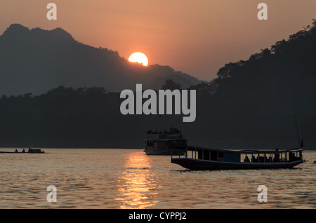 Bateaux du Mékong au coucher du soleil Luang Prabang Laos // LUANG PRABANG, Laos — le fleuve Mékong coule devant cette ville classée au patrimoine mondial de l'UNESCO tandis que des bateaux traditionnels en bois traversent la voie navigable au loin. La lumière dorée filtre à travers la brume atmosphérique au coucher du soleil, créant des silhouettes des montagnes environnantes. Luang Prabang se trouve au confluent du Mékong et du Nam Khan, dans le nord du Laos. La ville a servi de capitale royale du Royaume du Laos jusqu'en 1975 et reste un centre culturel important. Le transport fluvial traditionnel continue de jouer un rôle vital dans le commerce local et le Li quotidien Banque D'Images