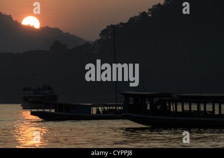 Bateaux du Mékong coucher de soleil Luang Prabang Laos // LUANG PRABANG, Laos — le fleuve Mékong coule devant cette ville classée au patrimoine mondial de l'UNESCO tandis que des bateaux traditionnels en bois traversent la voie navigable au loin. La lumière dorée filtre à travers la brume atmosphérique au coucher du soleil, créant des silhouettes des montagnes environnantes. Luang Prabang se trouve au confluent du Mékong et du Nam Khan, dans le nord du Laos. La ville a servi de capitale royale du Royaume du Laos jusqu'en 1975 et reste un centre culturel important. Le transport fluvial traditionnel continue de jouer un rôle vital dans le commerce local et la vie quotidienne Banque D'Images