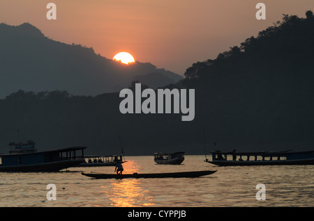 Bateaux traditionnels du Mékong Luang Prabang Laos // LUANG PRABANG, Laos — le fleuve Mékong coule devant cette ville classée au patrimoine mondial de l'UNESCO tandis que des bateaux traditionnels en bois traversent la voie navigable au loin. La lumière dorée filtre à travers la brume atmosphérique au coucher du soleil, créant des silhouettes des montagnes environnantes. Luang Prabang se trouve au confluent du Mékong et du Nam Khan, dans le nord du Laos. La ville a servi de capitale royale du Royaume du Laos jusqu'en 1975 et reste un centre culturel important. Le transport fluvial traditionnel continue de jouer un rôle vital dans le commerce local et quotidien Banque D'Images