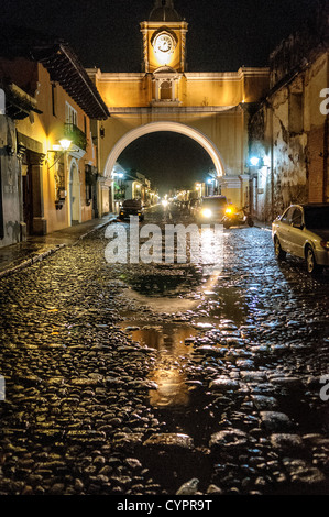 Santa Catalina Arch Night Antigua Guatemala / ANTIGUA GUATEMALA, Guatemala — L'arche sur la rue du couvent de Santa Catalina est illuminée la nuit, avec l'eau de la pluie récente reflétant les lumières sur la rue pavée. La tour de l'horloge emblématique au-dessus de l'arche se tient en évidence, tandis que le trottoir humide crée un chemin chatoyant pour les quelques véhicules qui traversent cette ville historique. Banque D'Images