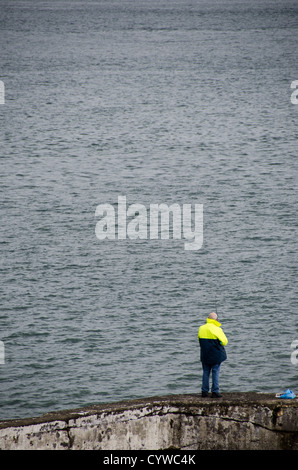 BEAUMARIS, pays de Galles — Un pêcheur solitaire jette sa ligne d'un vieux mur de pierre le long du front de mer de Beaumaris sur l'île d'Anglesey. La scène paisible capture l'attrait intemporel de la pêche à la ligne en mer avec pour toile de fond le détroit de Menai, avec les montagnes de Snowdonia visibles au loin. Cette image résume le charme tranquille et la beauté naturelle qui attirent les pêcheurs et les touristes dans cette pittoresque ville côtière galloise. Banque D'Images