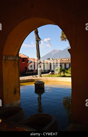 Volcan public source d'eau vue Antigua Guatemala // ANTIGUA, Guatemala — Une source d'eau publique ou une zone de lavage se dresse le long d'une rue pavée à Antigua, Guatemala, avec un volcan visible en arrière-plan émettant un panache de fumée. La structure en pierre, probablement l'une des fontaines publiques historiques de la ville ou « pilas » qui servaient autrefois de sources d'eau communautaires, reflète l'infrastructure coloniale de l'ancienne capitale. Antigua, site classé au patrimoine mondial de l'UNESCO depuis 1979, est connue pour son architecture coloniale espagnole bien préservée et est entourée de trois volcans, dont le Volcán de Agua Banque D'Images