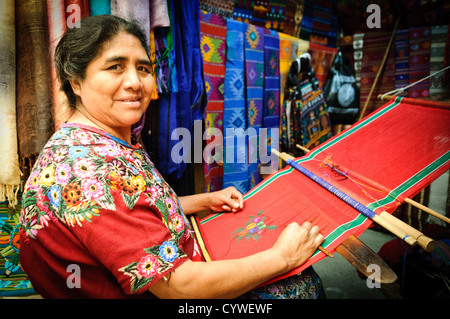 ANTIGUA GUATEMALA, Guatemala — Une femme maya fait la démonstration du tissage traditionnel de métier à tisser à sangle arrière, créant des textiles vibrants. Ses mains manipulent habilement des fils colorés, produisant des motifs complexes qui reflètent des techniques séculaires et un symbolisme culturel, tandis que des huipiles finies et d'autres articles tissés sont suspendus en arrière-plan. Banque D'Images