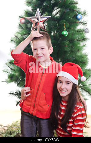 Young boy holding star sur le dessus de sa tête et sister wearing Santa hat in front of Christmas Tree, looking at camera, portrait Banque D'Images