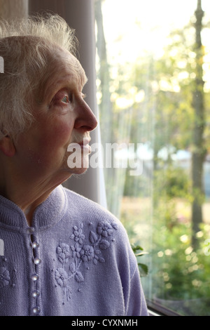 Une femme âgée regarde rêveusement hors de sa fenêtre, sentant ses pièges d'âge dans sa propriété. Banque D'Images