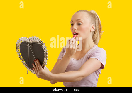 Belle jeune femme à la recherche de miroir tout en appliquant le rouge à lèvres sur fond jaune Banque D'Images