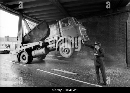 Un camion de saut coincé sous un pont semble être tenu par la main d'un policier alors qu'il contrôle la circulation 1985. La Grande-Bretagne des années 1980 route bloquée accident policier trafic collision camion coincé faible hauteur de pont Banque D'Images