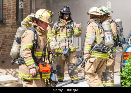 Pompiers américains équipe de secours pompiers devant un bâtiment en feu, discutant de stratégie tactique. Équipage diversifié caucasien, afro-américain. Banque D'Images