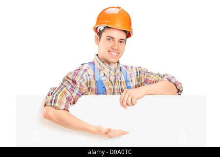 Young construction worker with helmet posant derrière un panneau et gesticulant isolé sur fond blanc Banque D'Images