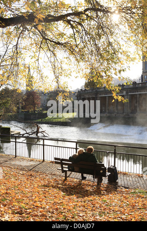 Deux amoureux sur un matin ensoleillé au début de l'hiver au bord de la rivière Avon, la ville de Bath avec les lames sur le banc de parc il y a Banque D'Images