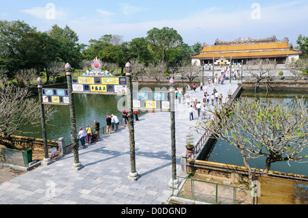 Pont d'entrée de la Cité Impériale Porte cérémonielle ornée Hué Vietnam | HUE, Vietnam — Les touristes traversent un pont de pierre à travers une porte cérémonielle ornée menant au Palais Thai Hoa au sein de la Cité Impériale. La passerelle centrale enjambant l'étang de la cour d'honneur du palais était historiquement réservée exclusivement aux empereurs Nguyễn ; les mandarins de la cour devaient utiliser les chemins latéraux lorsqu'ils approchaient de la salle du trône. Le Palais Thai Hoa, où 13 empereurs Nguyễn furent formellement intronisés, a survécu au tir d'artillerie qui a rasé une grande partie de la citadelle environnante pendant l'Offensive du Têt de 1968 et a depuis été Banque D'Images