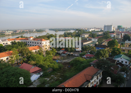 Rivière des Parfums et paysage urbain de Hué, Vietnam | HUÉ, Vietnam — Une vue élevée sur Hué montre la Rivière des Parfums sinuer à travers la ville, ses rives bordées de toits en tuiles rouges de l’ère coloniale et de gratte-ciel plus récents, dont l’hôtel Gerbera. La rivière sépare la citadelle impériale historique au nord des quartiers commerciaux au sud de l’eau, sous la brume humide typique du centre du Vietnam. Banque D'Images