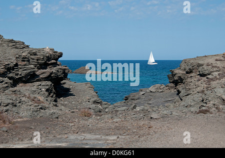 Voile blanc solitaire d'un yacht à voile dans les eaux bleu profond au large de la côte de Minorque Espagne à travers les rochers sur la rive Banque D'Images