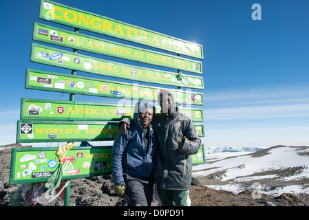 MONT KILIMANDJARO, Tanzanie — Un porteur pose avec l’un de ses clients à côté du panneau marquant le sommet du Mont Kilimandjaro, avec des nuages et de la glace bien au-dessous au loin. Banque D'Images