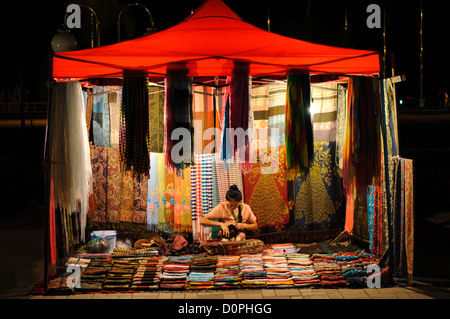 Vendeur de textile marché nocturne Quai Fa Ngum Vientiane Laos // VIENTIANE, Laos — Un vendeur exploite son étal textile sous une tente au marché nocturne du Quai Fa Ngum le long des rives du Mékong à Vientiane. Le marché nocturne du Quai Fa Ngum est une destination populaire en soirée où les vendeurs locaux vendent de l'artisanat, des textiles et des souvenirs aux habitants et aux touristes. Le quai Fa Ngum est la principale promenade riveraine de Vientiane, parallèle au Mékong en traversant le cœur de la capitale. La zone du front de mer a été développée comme un centre culturel et commercial, avec des restaurants Banque D'Images