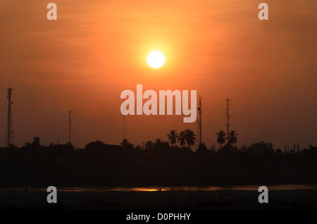 Couchant sur le fleuve Mékong à Vientiane Laos | VIENTIANE, Laos — Le soleil se couche derrière les tours de communication et les palmiers sur la rive thaïlandaise du Mékong, tel que vu depuis la capitale lao de l'autre côté de l'eau. Cette section marque la frontière fixée par le traité franco-siamois de 1893, qui a fait du fleuve la ligne de séparation entre l'Indochine française et le Siam et sépare encore aujourd'hui le Laos de la Thaïlande. Banque D'Images
