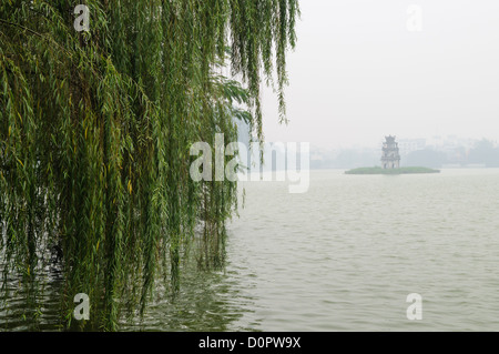 HANOI, Vietnam — Tour de la tortue (Thap Rua), sur une petite île au milieu du lac Hoan Kiem au cœur de Hanoi, est partiellement obscurcie par un épais brouillard matinal. Au premier plan, à gauche, se trouvent quelques-uns des arbres suspendus bas sur la rive du lac. Banque D'Images