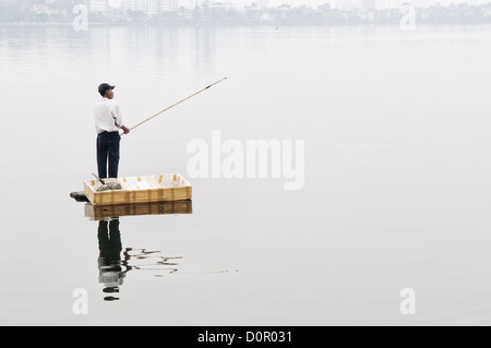 Pêcheur avec bâton de bambou Lac de l'Ouest Hanoi Vietnam // Un pêcheur se tient sur une petite plate-forme flottante pour lancer sa ligne d'une canne à pêche en bambou sur le Lac de l'Ouest (Ho Tay) à Hanoi, Vietnam. La brume épaisse obscurcit le rivage lointain. Une forte pollution dans le lac rend les poissons capturés là-bas de qualité douteuse. Banque D'Images
