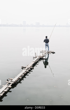 Pêcheur sur la jetée West Lake Hanoi Vietnam // Un pêcheur se tient sur une jetée étroite en bois pour jeter sa ligne sur le lac West (Ho Tay) à Hanoi, Vietnam. La brume épaisse obscurcit le rivage lointain. Comme la plupart des lacs de Hanoi, le lac de l'Ouest est fortement pollué, ce qui rend le poisson pêché là d'une qualité douteuse pour manger. Banque D'Images