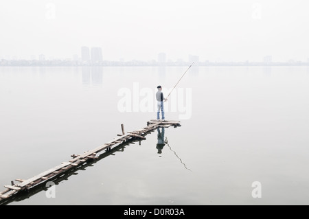 Pêcheur sur la jetée West Lake Hanoi Vietnam // Un pêcheur se tient sur une jetée étroite en bois pour lancer sa ligne sur le lac West (Ho Tay) à Hanoi, Vietnam, en utilisant une canne combinée avec une ligne séparée à la main plutôt qu'un rouleau fixe. La brume épaisse obscurcit le rivage lointain. Comme la plupart des lacs de Hanoi, le lac de l'Ouest est fortement pollué, ce qui rend le poisson pêché là d'une qualité douteuse pour manger. Banque D'Images