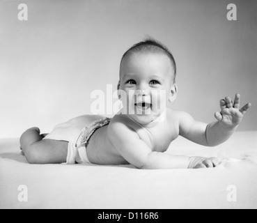 1960 SMILING BABY WEARING couche en tissu couché sur l'estomac à la caméra AU BRAS LEVÉ Banque D'Images