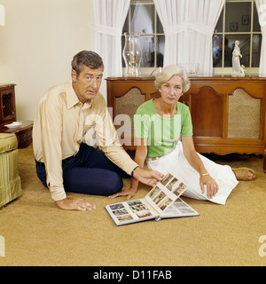 1970 COUPLE D'ÂGE MOYEN AVEC PLANCHER DE SÉANCE PHOTO ALBUM LOOKING AT CAMERA Banque D'Images