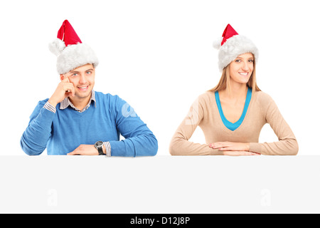 Smiling couple wearing santa hats et posant derrière un panneau vierge Banque D'Images