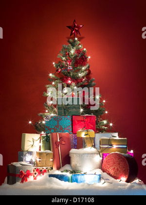 Arbre de Noël décoré magnifiquement entouré par un tas de cadeaux colorés couverts de neige sur fond rouge Banque D'Images