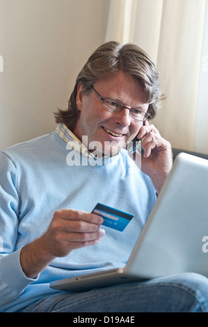 Homme mûr à la maison faire une carte de transaction financière en ligne à l'aide de son ordinateur portable et un smartphone iPhone Banque D'Images