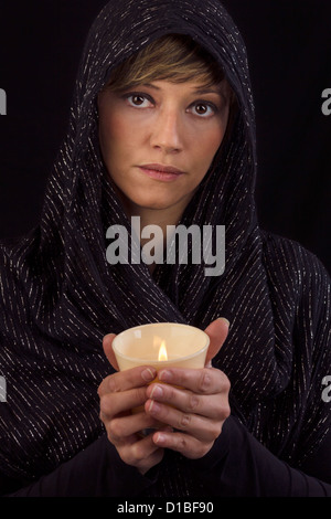 Belle jeune femme en cape noire tenant une bougie allumée dans ses mains et réfléchi à l'appareil photo. Portrait Studio Banque D'Images