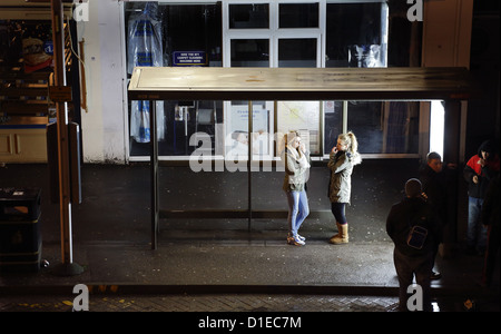 Deux jeunes femmes qui attendent à un arrêt d'autobus de nuit à Londres, Royaume-Uni. Un groupe de jeunes hommes sont à proximité. Banque D'Images