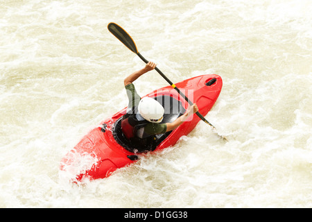 Une vue aérienne d'un kayakiste dans un kayak rouge vif manoeuvrant à travers des rapides turbulents d'eau vive. Le kayakiste, portant un casque blanc et un haut à manches longues vert foncé, pagaie activement avec un Banque D'Images