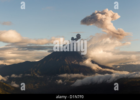 Une grande montagne volcanique est activement en éruption, libérant un panache de cendres foncées de son sommet qui s’élève dans un ciel partiellement nuageux. Les pentes accidentées et boisées du volcan sont partiellement entourées Banque D'Images