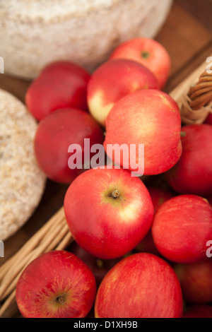 Pommes rouges s'échappant d'un panier en osier à côté du fromage sur une planche en bois Banque D'Images