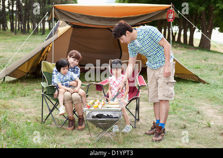 Cuisson tandis que le père d'autres membres de la famille assis sur chaises de camping Banque D'Images