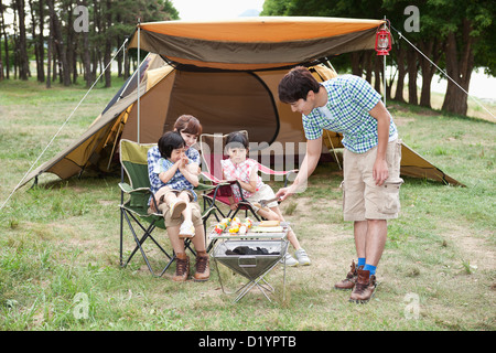 Cuisson tandis que le père d'autres membres de la famille assis sur chaises de camping Banque D'Images