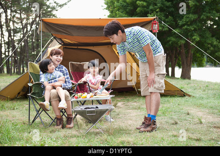 Cuisson tandis que le père d'autres membres de la famille assis sur chaises de camping Banque D'Images