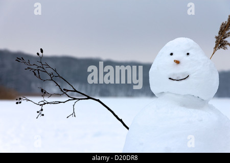 A smiling happy snowman soulève la brindille qu'il a pour un bras en guise de salutation avant de l'arrière-plan d'un lac gelé et la forêt. Banque D'Images