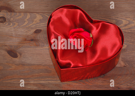 Une rose rouge dans une boîte en forme de coeur rouge avec doublure satin contre un arrière-plan en bois rustique. Banque D'Images