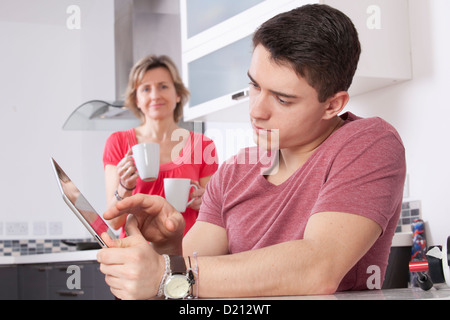 Jeune homme à l'aide d'une tablette numérique à la recherche à l'écran ou moniteur. Situé dans une cuisine moderne une femme tenant deux mugs sourit. Banque D'Images