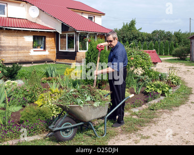 Jardinier principal près de flower bed avec fourche et brouette avec les mauvaises herbes Banque D'Images