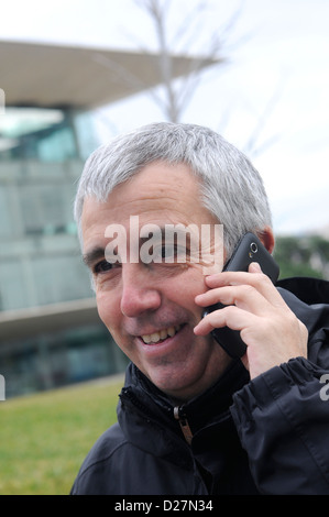 La photographie de rue moyen age man smiling, écoute mobile smartphone avec des cheveux blancs et manteau noir, 50 ans rides Banque D'Images