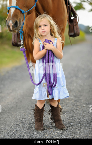 Petite blonde jeune fille avec son gros cheval sur un chemin de campagne, pose assurée avec regard soutenu, Soleil bleu robe et hautes. Banque D'Images