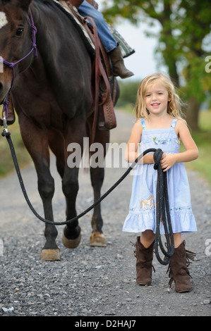 Petite blonde cowgirl menant son grand cheval vers le bas une route de campagne, souriant et portant robe bleue et bottes western. Banque D'Images