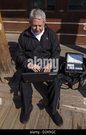 Homme senior assis sur une marche en bois utilisant le point d'accès WiFi local créant ainsi son propre bureau extérieur dans le centre-ville de Dubois, Wyoming, Etats-Unis. Banque D'Images