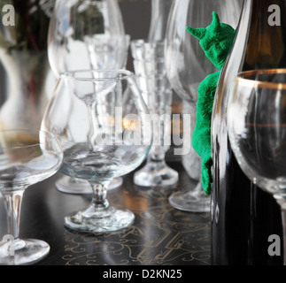 un petit diable vert, de l'argile à modeler, sort de derrière une bouteille de vin. Une variété de verres à boire sont étalées sur une table incrusté. Banque D'Images